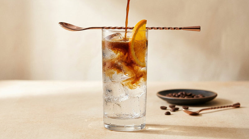 Espresso turnat peste dosul unei linguri într-un pahar înalt cu tonic și gheață, cu straturile vizibile și o felie de portocană pe margine