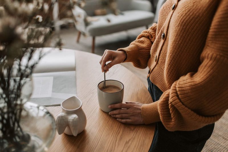 Cană de cafea cu lapte amestecată cu lingurița, pe o masă de lemn, lumină caldă de toamnă